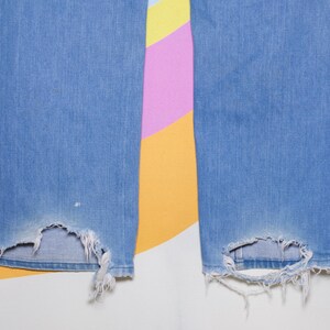 Puede incluir: Primer plano de unos vaqueros de mezclilla azul claro con dobladillos rotos y desgastados. Los vaqueros tienen un dise&ntilde;o cl&aacute;sico de cinco bolsillos y un corte recto. Los bordes deshilachados de las zonas rotas a&ntilde;aden un aspecto vintage y desgastado. El fondo es un patr&oacute;n abstracto colorido.