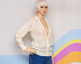 May include: Cream-colored, open-weave cardigan with long sleeves and a button-down front. The sweater has a delicate, lacy pattern and ribbed cuffs and hem. The garment is displayed on a mannequin.