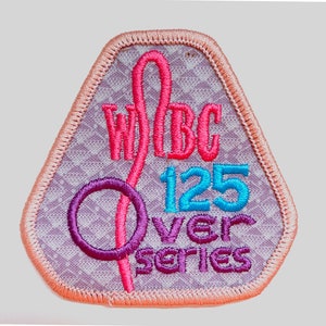 May include: Embroidered patch with a light purple background and a peach-colored border. The patch features the text "WBC 125 Over Series" in pink, blue, and purple thread. The design includes a stylized ribbon and circle.