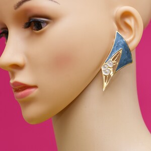 May include: A gold-toned, geometric earring with a blue enamel section and white floral accents. The earring is a bold, asymmetrical design, showcasing a modern aesthetic. The earring is displayed on a mannequin.