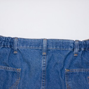 Puede incluir: Primer plano de la cintura y los bolsillos traseros de unos pantalones cortos vaqueros azules. Los pantalones cortos tienen una cintura el&aacute;stica, trabillas y dos bolsillos traseros con costuras decorativas. El denim es de lavado medio.