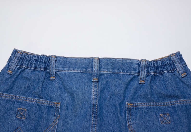 Puede incluir: Primer plano de la cintura y los bolsillos traseros de unos pantalones cortos vaqueros azules. Los pantalones cortos tienen una cintura el&aacute;stica, trabillas y dos bolsillos traseros con costuras decorativas. El denim es de lavado medio.