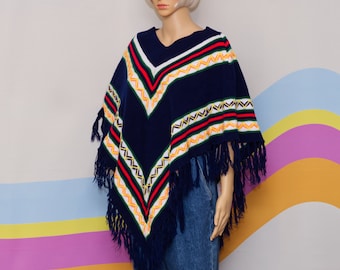 May include: A navy blue knitted poncho with a V-neck and fringe detailing. The poncho features a geometric pattern in red, white, yellow, and green. The garment is displayed on a mannequin.
