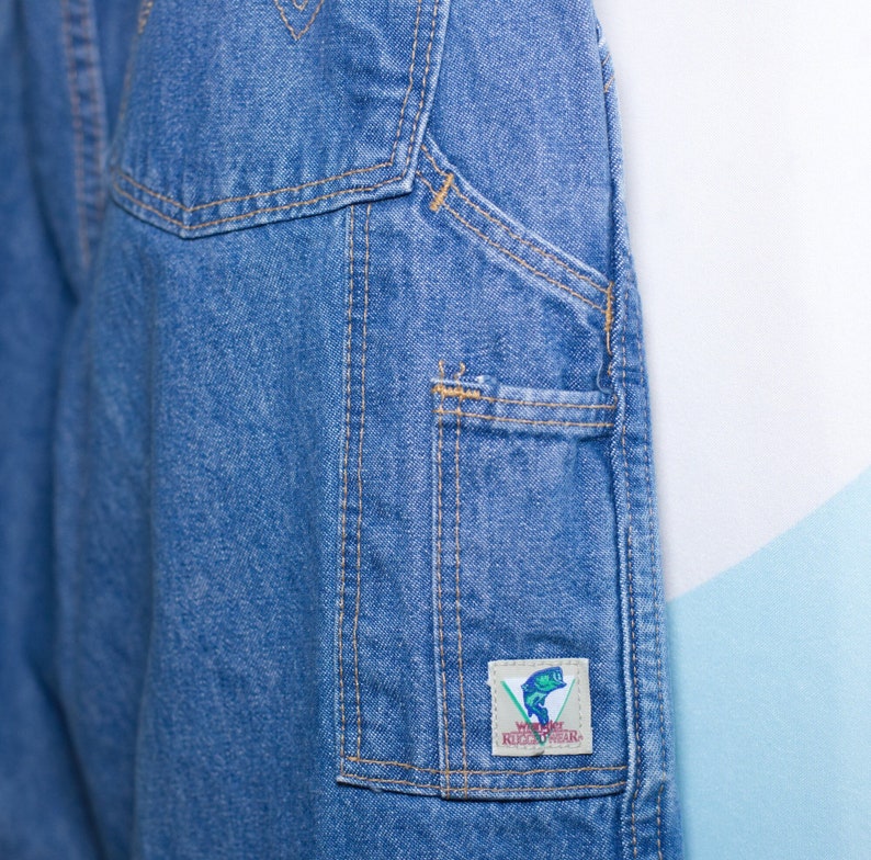 Puede incluir: Primer plano de un pantal&oacute;n de trabajo de mezclilla azul con m&uacute;ltiples bolsillos y costuras visibles. Un peque&ntilde;o parche rectangular en el bolsillo presenta un gr&aacute;fico de un pez y el texto "WEAR KICKWEAR".