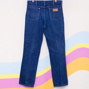 May include: Dark blue denim Wrangler jeans hanging against a colorful background. The jeans have classic five-pocket styling, a brown leather patch, and copper-colored rivets. The back pockets feature the signature Wrangler stitching.