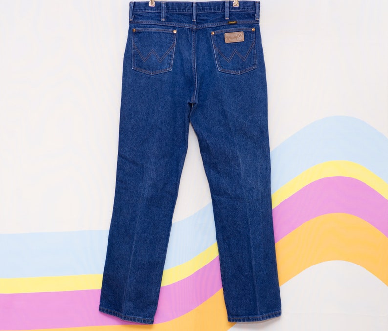 May include: Dark blue denim Wrangler jeans hanging against a colorful background. The jeans have classic five-pocket styling, a brown leather patch, and copper-colored rivets. The back pockets feature the signature Wrangler stitching.