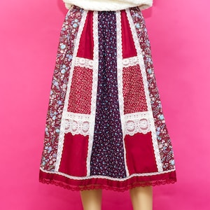May include: A patchwork skirt in a variety of patterns and colours. The skirt features panels of burgundy, red, and navy blue fabric, with white lace trim. The hemline has a delicate lace border, creating a vintage aesthetic. The skirt is approximately 76 cm in length.