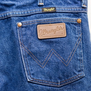 May include: Close-up of the back pocket of blue denim Wrangler jeans. The pocket features a brown leather patch with the Wrangler logo and is stitched with a contrasting tan thread. The jeans have a classic five-pocket design.