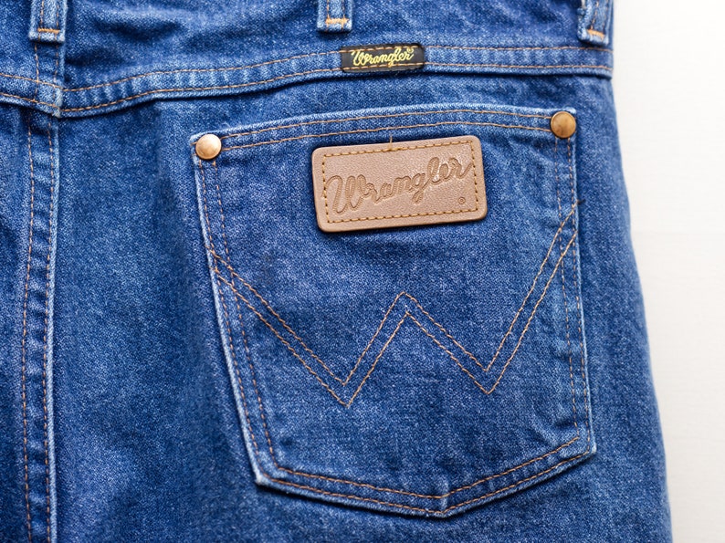 May include: Close-up of the back pocket of blue denim Wrangler jeans. The pocket features a brown leather patch with the Wrangler logo and is stitched with a contrasting tan thread. The jeans have a classic five-pocket design.