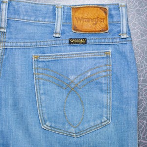 Puede incluir: Vaqueros de mezclilla azul claro con un parche de cuero Wrangler y una peque&ntilde;a etiqueta Wrangler. El bolsillo trasero presenta un dise&ntilde;o de costura decorativo. Vaqueros de estilo cl&aacute;sico.