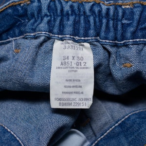 Puede incluir: Primer plano de unos vaqueros azules con una etiqueta. La etiqueta muestra la talla 34 x 30 pulgadas, y el material es 100% algod&oacute;n. La etiqueta tambi&eacute;n incluye el texto "Made in USA".