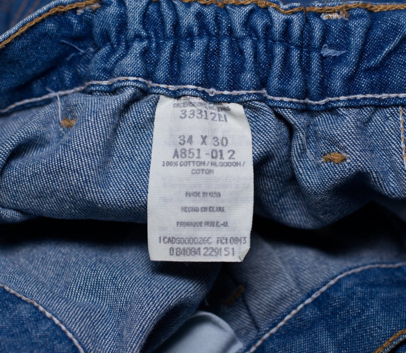 Puede incluir: Primer plano de unos vaqueros azules con una etiqueta. La etiqueta muestra la talla 34 x 30 pulgadas, y el material es 100% algod&oacute;n. La etiqueta tambi&eacute;n incluye el texto "Made in USA".