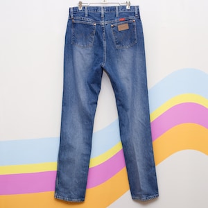 May include: Blue denim jeans hanging on a metal hanger. The jeans feature a classic five-pocket design, a brown leather patch with a red logo, and a straight leg cut. The jeans are a medium wash.