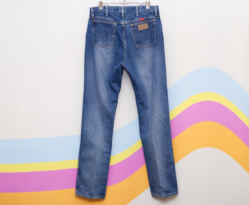 May include: Blue denim jeans hanging on a metal hanger. The jeans feature a classic five-pocket design, a brown leather patch with a red logo, and a straight leg cut. The jeans are a medium wash.