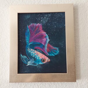 Puede incluir: Obra de arte enmarcada con un vibrante pez betta. El pez tiene un cuerpo azul y rosa con una cola grande y fluida en tonos de azul, rosa y rojo. El fondo es oscuro y el marco es de color oro claro.