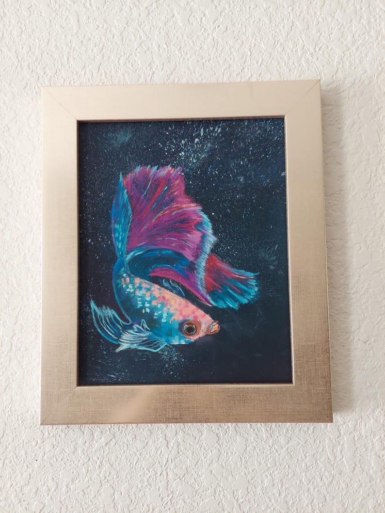 Puede incluir: Obra de arte enmarcada con un vibrante pez betta. El pez tiene un cuerpo azul y rosa con una cola grande y fluida en tonos de azul, rosa y rojo. El fondo es oscuro y el marco es de color oro claro.