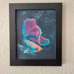 Puede incluir: Obra de arte enmarcada con un pez betta vibrante. El pez tiene un cuerpo turquesa y aletas rosas y azules, sobre un fondo azul oscuro. La obra est&aacute; en un marco negro, adecuada para exhibir en la pared.