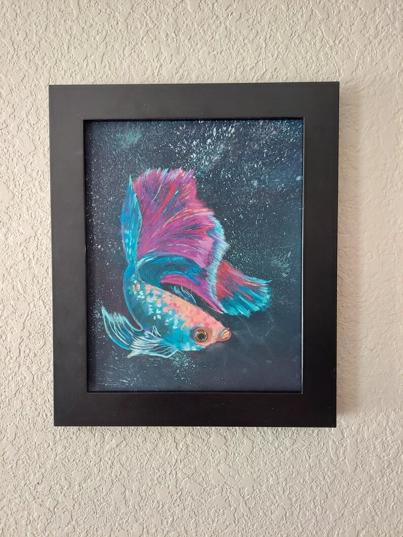 Puede incluir: Obra de arte enmarcada con un pez betta vibrante. El pez tiene un cuerpo turquesa y aletas rosas y azules, sobre un fondo azul oscuro. La obra est&aacute; en un marco negro, adecuada para exhibir en la pared.