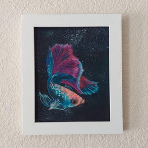 Puede incluir: Pintura enmarcada de un pez luchador. El pez tiene un cuerpo azul y naranja con una cola grande y fluida en tonos de azul y magenta. El fondo es oscuro con motas blancas y el marco es blanco.