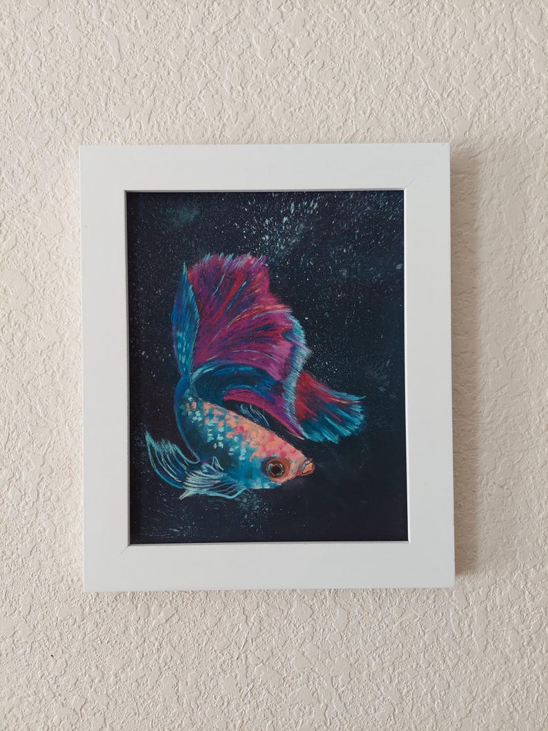Puede incluir: Pintura enmarcada de un pez luchador. El pez tiene un cuerpo azul y naranja con una cola grande y fluida en tonos de azul y magenta. El fondo es oscuro con motas blancas y el marco es blanco.