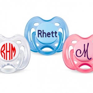 May include: Three baby pacifiers in clear, blue, and pink. The clear pacifier has a red monogram, the blue one says "Rhett", and the pink one has a cursive "M". All have a rounded shield and a ring handle.