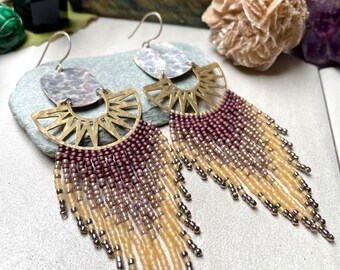 Może przedstawiać: Para ręcznie robionych kolczyków w stylu boho. Kolczyki posiadają srebrny dysk, złoty wzór słońca i frędzle z koralików w odcieniach bordowego, beżowego i srebrnego. Kolczyki mają zapięcie na haczyk.