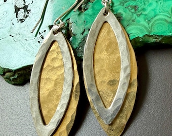 Może przedstawiać: Para ręcznie robionych kolczyków o warstwowym wzorze. Każdy kolczyk ma złotą podstawę w kształcie liścia, na której nałożona jest srebrna, owalna ramka. Kolczyki mają młotkowaną fakturę i są zawieszone na srebrnych haczykach.