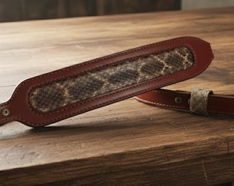 May include: A brown leather rifle sling with a snakeskin-patterned center panel. The sling is made of high-quality leather and features a buckle for adjustment. The design is classic and suitable for hunting or sporting rifles.