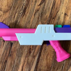 May include: A brightly coloured toy gun. The barrel is pink, with a white body and a pink handle. The top has purple, green, and blue accents. The toy is made of plastic and sits on a wooden surface.