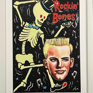 May include: A framed art print with a black background featuring a yellow skeleton wearing a hat and a blue bow tie, and a man with blonde hair. The words "Rockin' Bones" are in red. Musical notes and stars are scattered around.