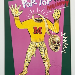 May include: A framed Pop-Top The Mummy artwork. The illustration features a yellow mummy figure with a detached head, wearing a yellow sweater with a large red "M". The background is pink with white text and a green border.