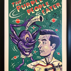 May include: A framed art print titled THE PURPLE PEOPLE EATER featuring a purple one-eyed creature with a horn and a person playing an acoustic guitar against a green space background with red planets and musical notes. The print is signed 2/10.
