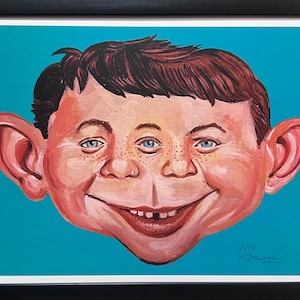 May include: A framed art print featuring a stylized, caricature-style portrait of a person with brown hair, large ears, and three blue eyes. The face has a wide, toothy grin and freckles, set against a solid teal background with a signature at the bottom.