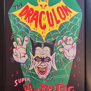 May include: Framed artwork featuring a vintage-style illustration of a vampire character. The artwork includes the text "NEW Monster" and "THE DRACULON" in red, with "SUPER Horrific" at the bottom. The background is green with a spiderweb design.