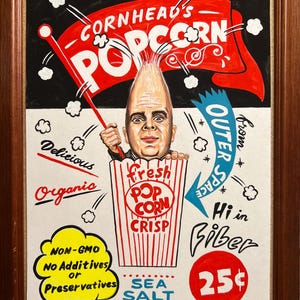 May include: A framed vintage-style popcorn advertisement. The artwork features a man with a popcorn-shaped head holding a flag that reads "Cornhead's Popcorn." Text includes "Delicious," "Organic," "Non-GMO," and "Sea Salt." A red circle displays "25¢."