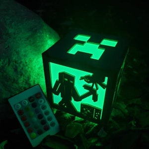 May include: A Minecraft-themed cube lamp with a green glow. The lamp features cut-out designs of characters and symbols from the game. A remote control with color options sits nearby. The lamp is made of dark wood.