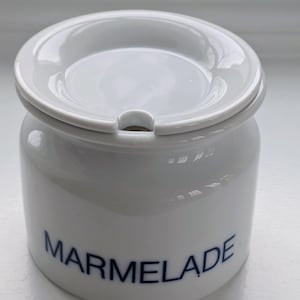 Puede incluir: Tarro de mermelada de cerámica blanca con tapa a juego. El tarro tiene forma redondeada con la palabra "MARMELADE" en letras azules. La tapa tiene una pequeña abertura para una cuchara.