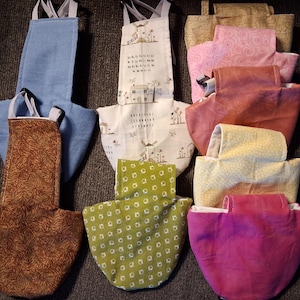 May include: A collection of handmade fabric aprons in various colors and patterns. The aprons include a light blue, brown leaf print, white with a windmill design, green with white dots, pink swirl, and a yellow honeycomb pattern.