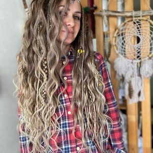 May include: A woman with long, wavy, blonde hair with braids and beads. She is wearing a red and black plaid shirt.