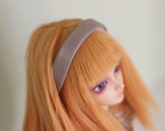 May include: A doll with orange hair and a taupe headband. The headband is made of a textured material and sits across the doll's forehead. The doll has blue eyes and a pale complexion. The background is a soft, neutral color.