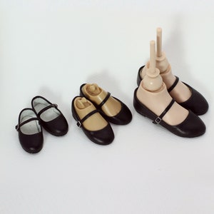 May include: Four pairs of black leather doll shoes with a strap and buckle. The shoes are on a white background.