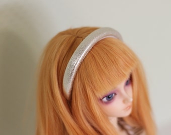 May include: A silver glitter headband sits atop a doll's orange hair. The headband is a simple, rounded design, and the glitter catches the light. The doll has blue eyes and a pale complexion.