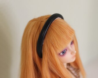 May include: A black headband with a rounded top is worn on a doll with orange hair. The doll has pale skin, purple eye shadow, and blue eyes. The headband is made of a smooth, dark material.