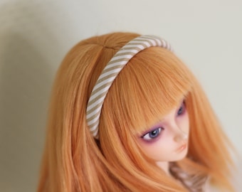 May include: A doll with orange hair and blue eyes wearing a striped headband. The headband features a pattern of beige and white stripes. The doll's hair is styled with a side part.