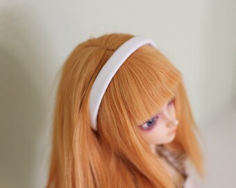 May include: A white headband sits atop a doll's head with long, orange hair. The headband is a simple, rounded band. The doll has blue eyes and a pale complexion. The background is a soft, neutral color.
