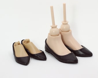 May include: Two pairs of miniature doll shoes. One pair is black and tan ballet flats, and the other is a pair of black flats attached to doll feet with tan skin. The background is white.