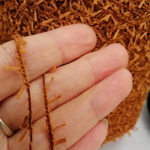 May include: Close-up of a brown yarn with fringe. The yarn is held in a hand.