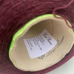 Op de afbeelding: Een close-up van een bruine bol garen met een label waarop "Loro Piana" staat en details zoals "Cod. C282 54353", "Nm. CASHMERE 2/27", "100% WS", "Des. 49 DANDY UN =4R01", "TESS LGQ319", "PE", "Pita-Lot 243708" en "Data-Date 17/11/2022" vermeld staan.