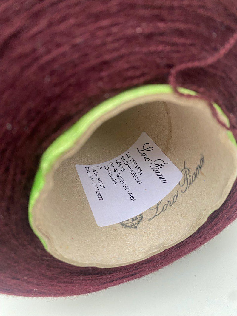 Op de afbeelding: Een close-up van een bruine bol garen met een label waarop "Loro Piana" staat en details zoals "Cod. C282 54353", "Nm. CASHMERE 2/27", "100% WS", "Des. 49 DANDY UN =4R01", "TESS LGQ319", "PE", "Pita-Lot 243708" en "Data-Date 17/11/2022" vermeld staan.