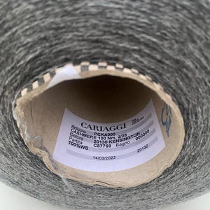May include: A close-up of a gray yarn ball with a white label that reads "CARIAGGI" and includes details like "PCKA000", "CASHMERE 100 Nm. 2/28", "20130 KENSINGTON", "C87769", "100%WS", and "14/03/2023".